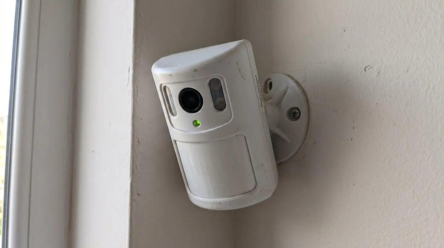 Détecteur de mouvement blanc avec objectif caméra intégré monté dans l'angle d'un mur intérieur, capteurs infrarouges et ouverture caméra visibles en gros plan